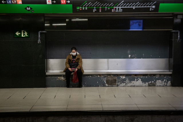 Una mujer protegida con mascarilla espera sentada en un banco del andén a que llegue el metro, durante el noveno día laborable desde que se decretó el estado de alarma en el país a consecuencia del coronavirus, en Barcelona/Catalunya (España) a 26 de marz
