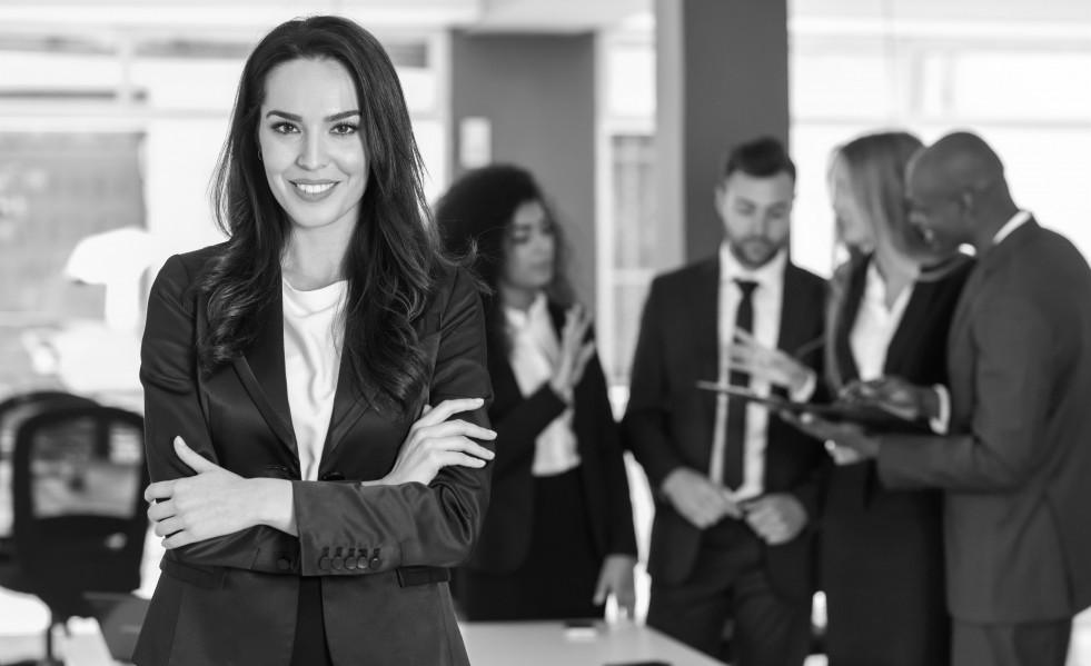 Archivo - Líder empresaria mirando a cámara en la oficina moderna con empresarios multiétnicos que trabajan en segundo plano